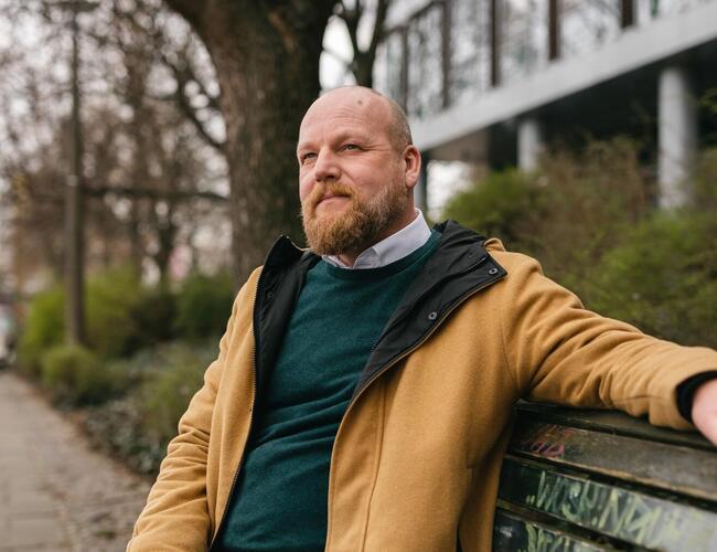 Matthias Krahe, Abteilungsleiter Humanistische Lebenskunde im Humanistischen Verband Berlin-Brandenburg, sitze auf einer Bank, wobei sein linker Arm oben auf der Lehne ruht.