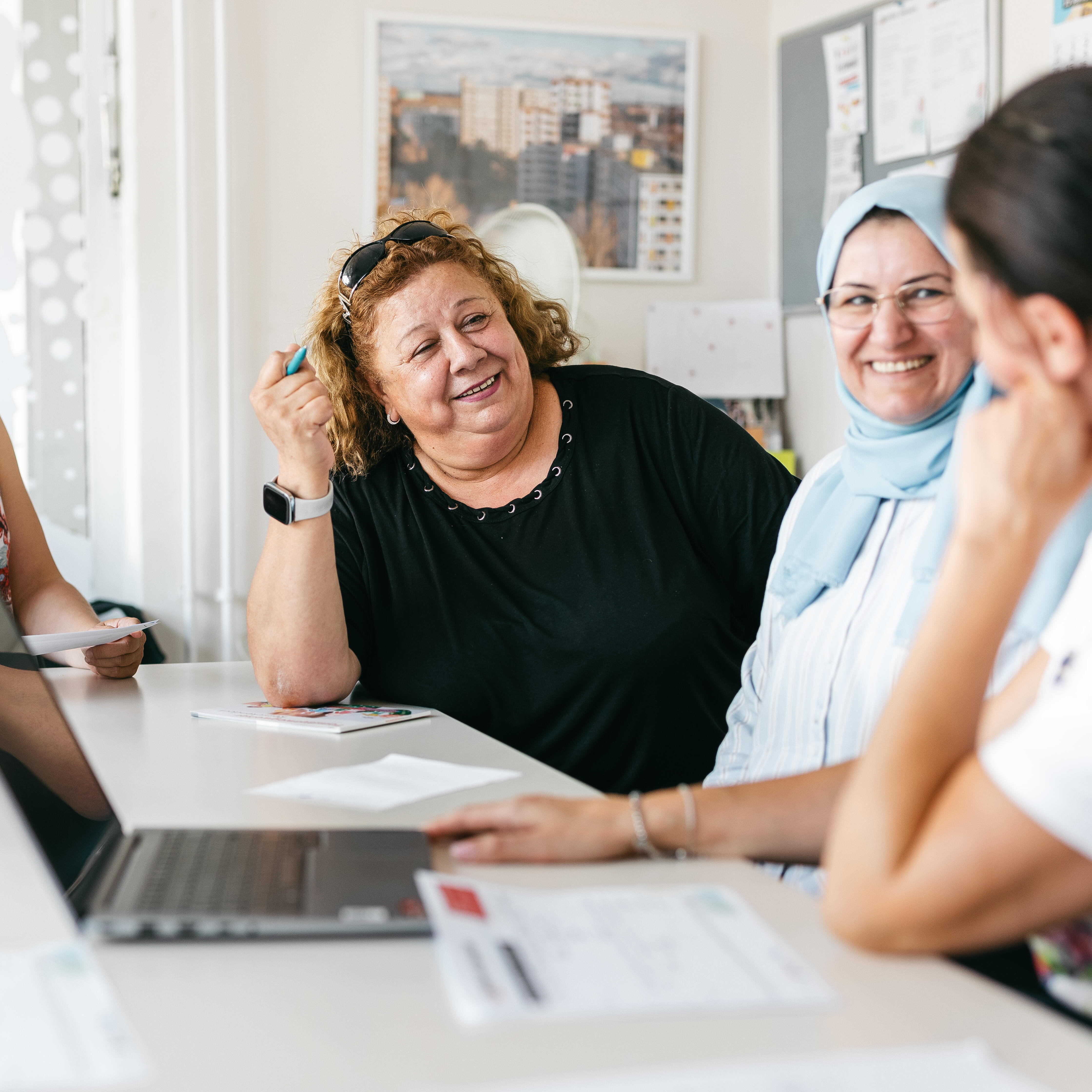 Zu sehen sind vier Frauen, die an einem weißen Tisch sitzen. Drei von ihnen sehen wir von vorne, sie schauen auf die Frau ganz rechts, die wir nur verschwommen erkennen können. Sie lächeln sich an, auf dem Tisch steht ein Laptop.