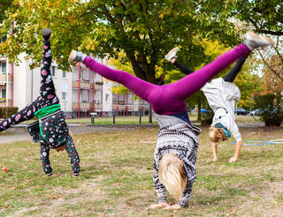 M*ZW Teltow Sport und Bewegung Zu sehen sind drei junge Personen auf einer Wiese, die jeweils einen Radschlag machen. Sie haben die Beine in der Luft und stützen sich auf ihren Armen. Die Person vorne trägt eine knallige, lila Hose und eine gemusterte Jacke.