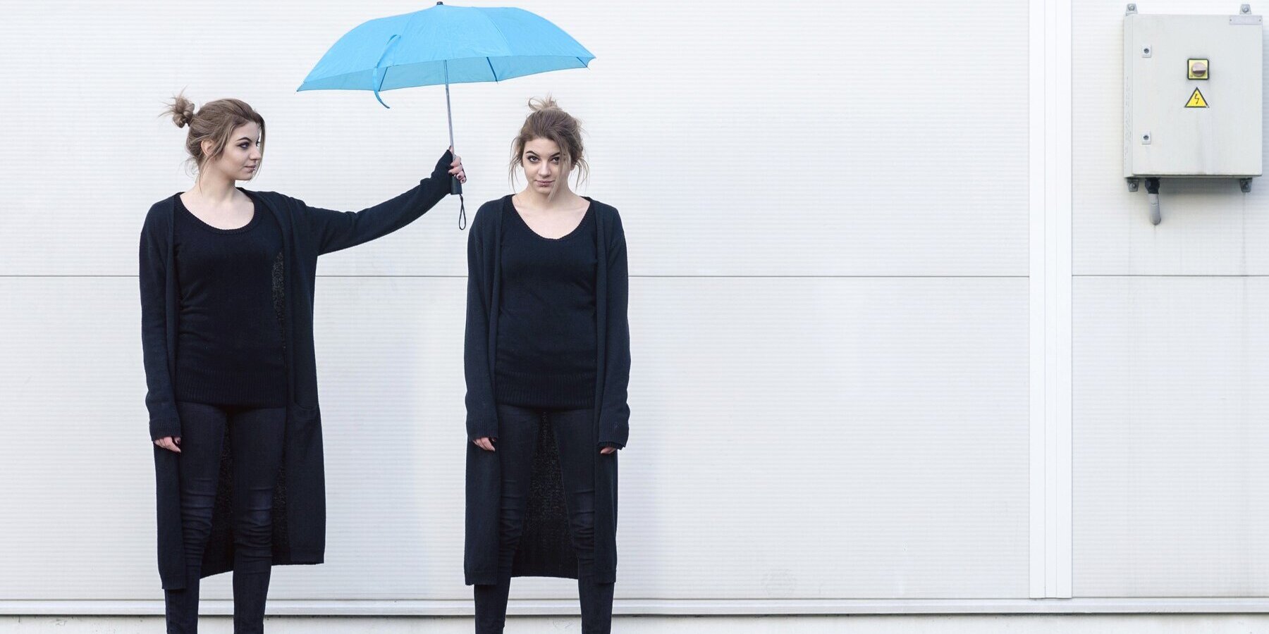 Eine junge Frau in schwarzer Kleidung hält einen blauen Regenschirm über den Kopf einer weiteren jungen Frau, die genauso aussieht wie die Frau, die den Regenschirm hält.