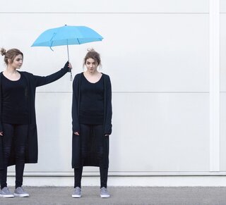 Kontakt- und Informationsstelle für Selbsthilfe in Berlin-Pankow: Peer-Beratung Eine junge Frau in schwarzer Kleidung hält einen blauen Regenschirm über den Kopf einer weiteren jungen Frau, die genauso aussieht wie die Frau, die den Regenschirm hält.