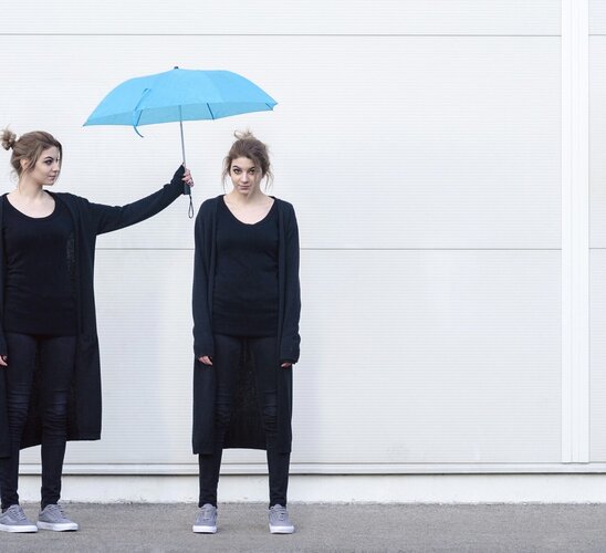 Eine junge Frau in schwarzer Kleidung hält einen blauen Regenschirm über den Kopf einer weiteren jungen Frau, die genauso aussieht wie die Frau, die den Regenschirm hält.