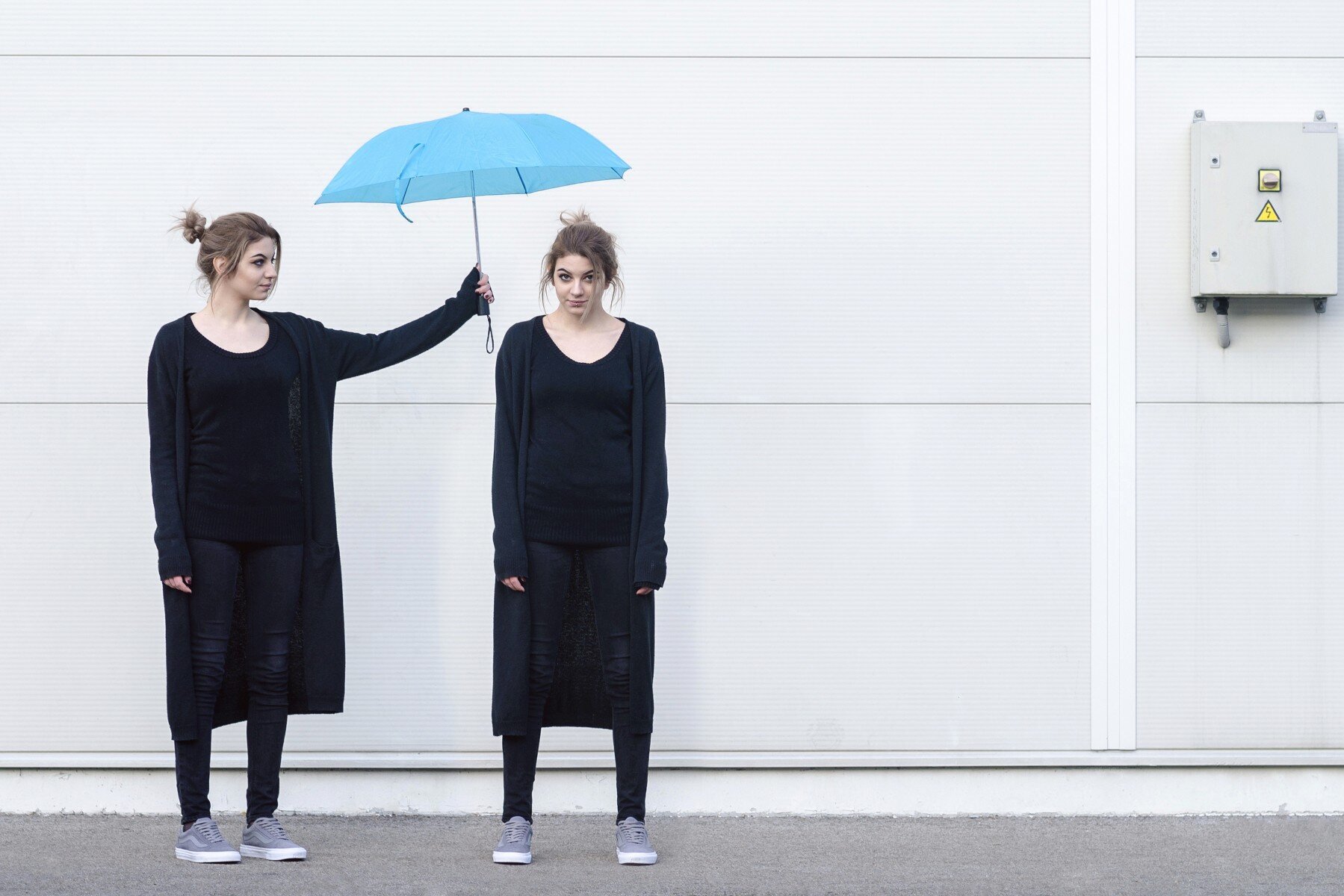 Eine junge Frau in schwarzer Kleidung hält einen blauen Regenschirm über den Kopf einer weiteren jungen Frau, die genauso aussieht wie die Frau, die den Regenschirm hält.