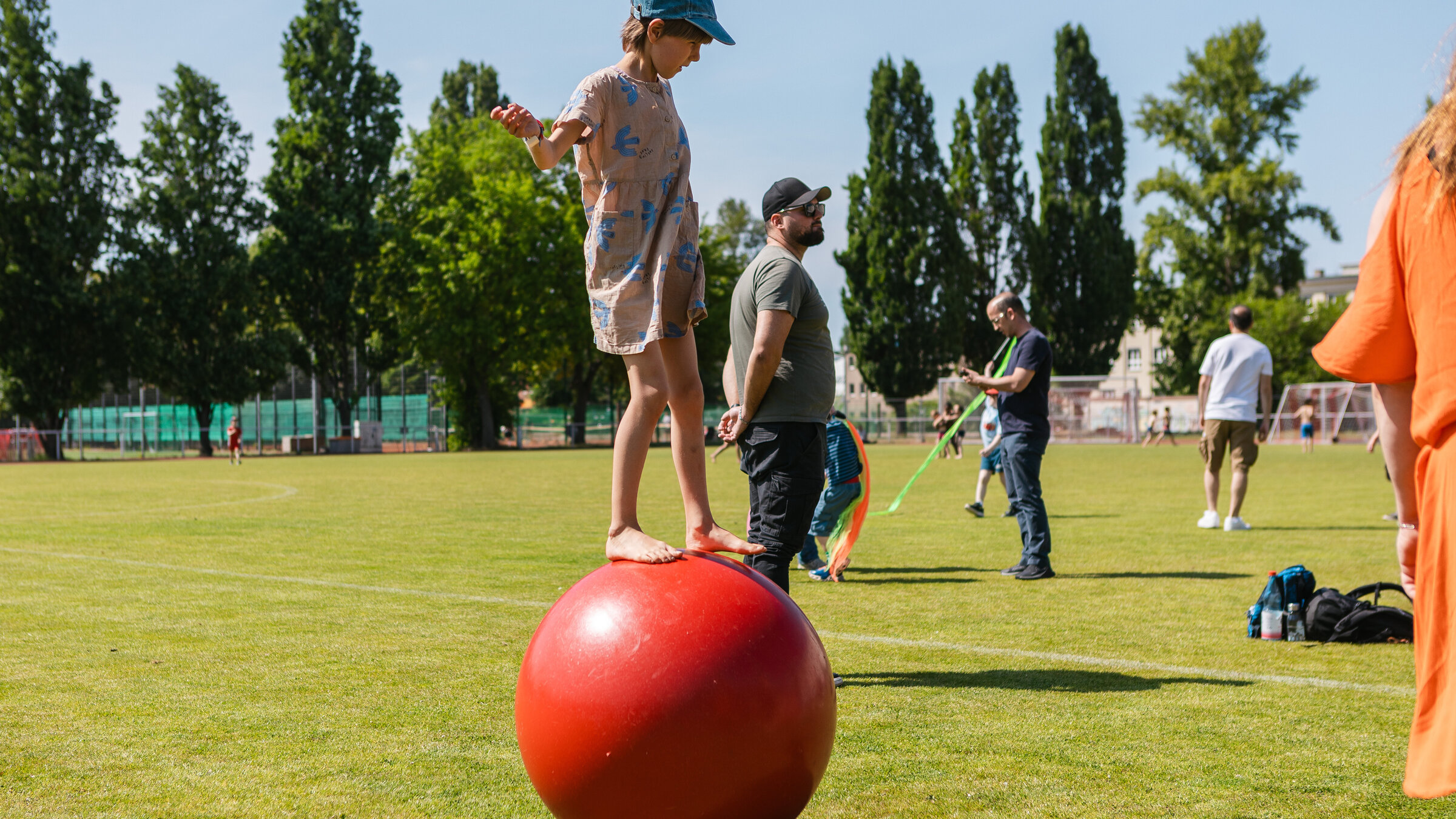 Ein kleines Mädchen balanciert barfuß auf einem großen roten Ball auf einer grünen Wiese.