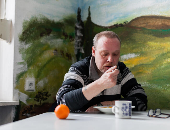 Ein Mann sitzt in einer Ecke an einem Tisch und isst gerade von einem Löffel. Vor ihm steht ein gefüllter Teller, eine Tasse, eine Brille und eine Mandarine.