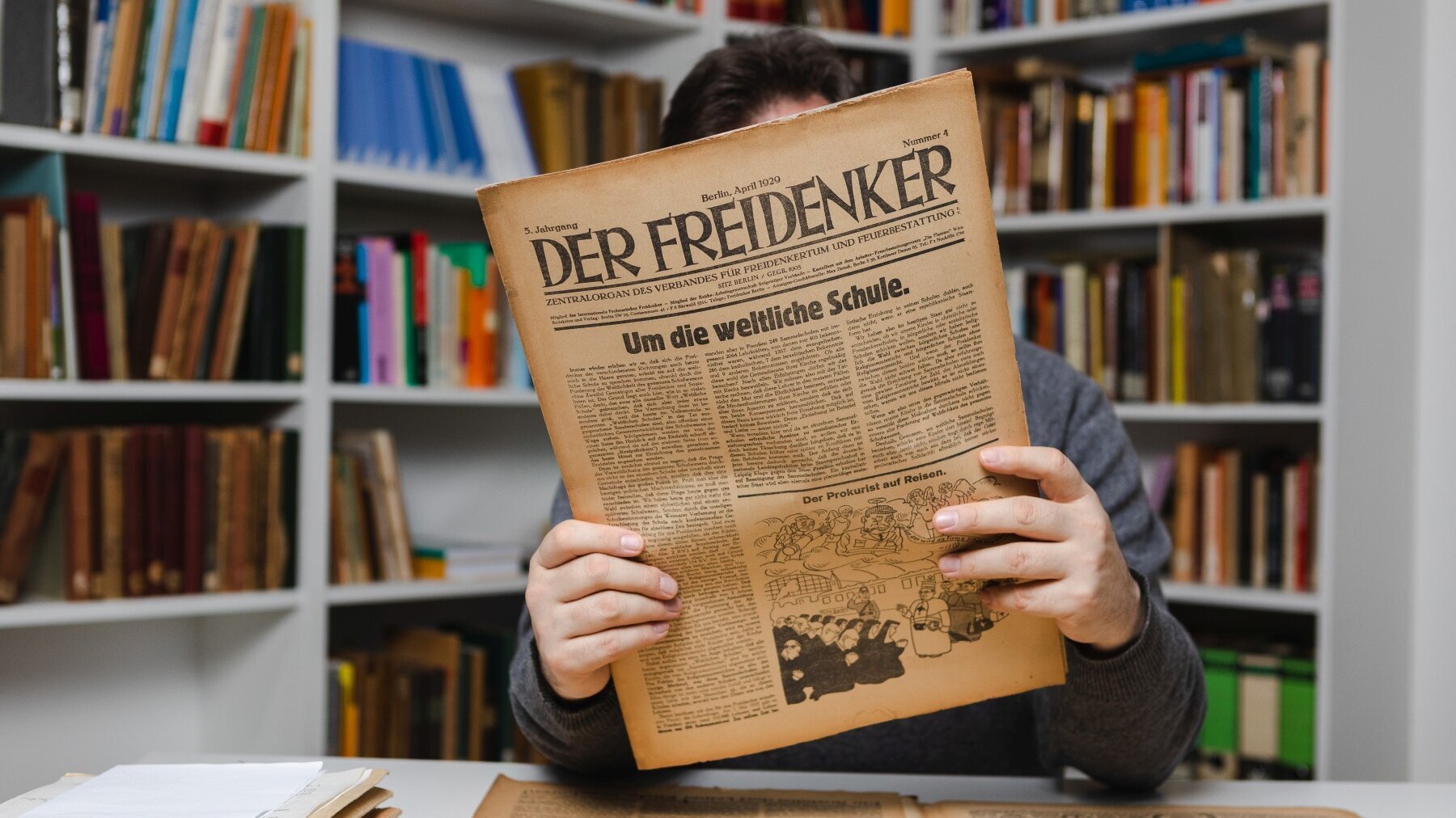 An einem Tisch in einem Raum mit Regalen voller Aktenordner und Bücher sitzt eine Person. Ihr Gesicht wird durch eine historische Zeitschrift verdeckt, die die Person in ihren Händen hält.