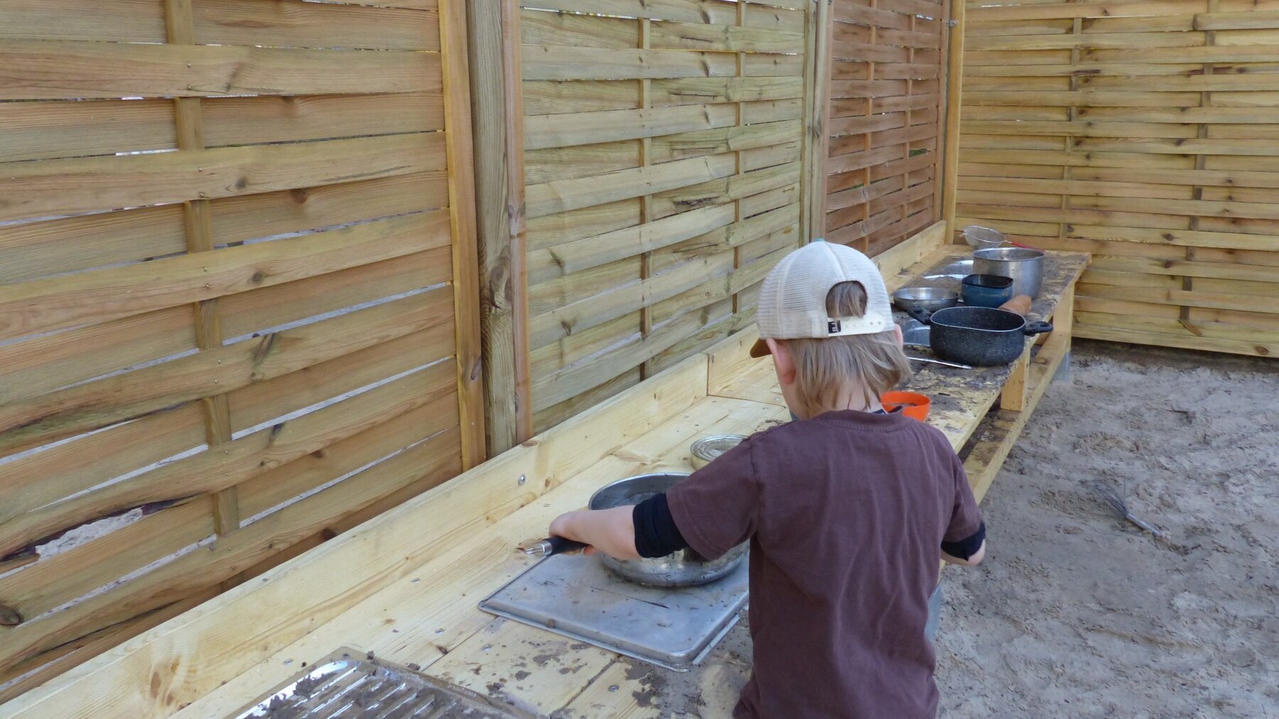 Ein Junge steht an einer Holzbank mit Metallplatten und Kochtöpfen darauf und kocht mit Sand.