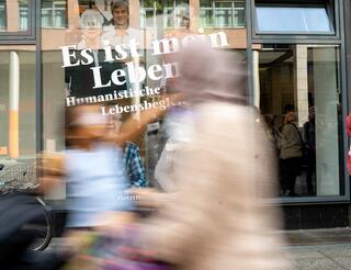 Haus des HUMANISMUS Leipziger Straße Eröffnung Menschen spiegeln sich in einem Schaufenster auf dem steht: "Es ist mein Leben".