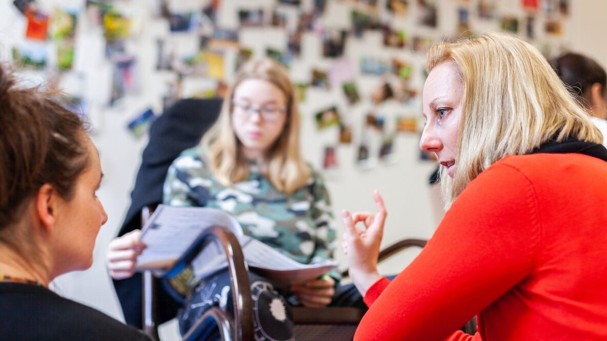 Eine Studierende, die etwas auf Papier liest, sitzt im Hintergrund auf einem Stuhl und ist verschwommen zu sehen. Davor hockt rechts eine Dozentin und links eine Studierende, der die Dozentin etwas erklärt.