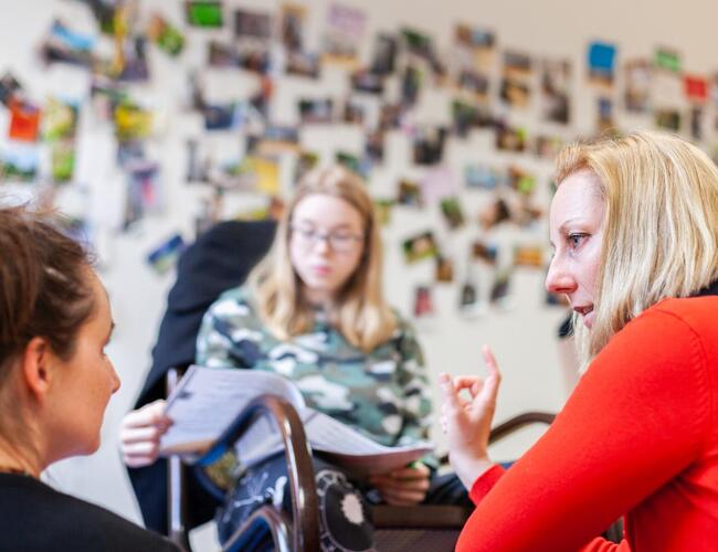 Eine Studierende, die etwas auf Papier liest, sitzt im Hintergrund auf einem Stuhl und ist verschwommen zu sehen. Davor hockt rechts eine Dozentin und links eine Studierende, der die Dozentin etwas erklärt.
