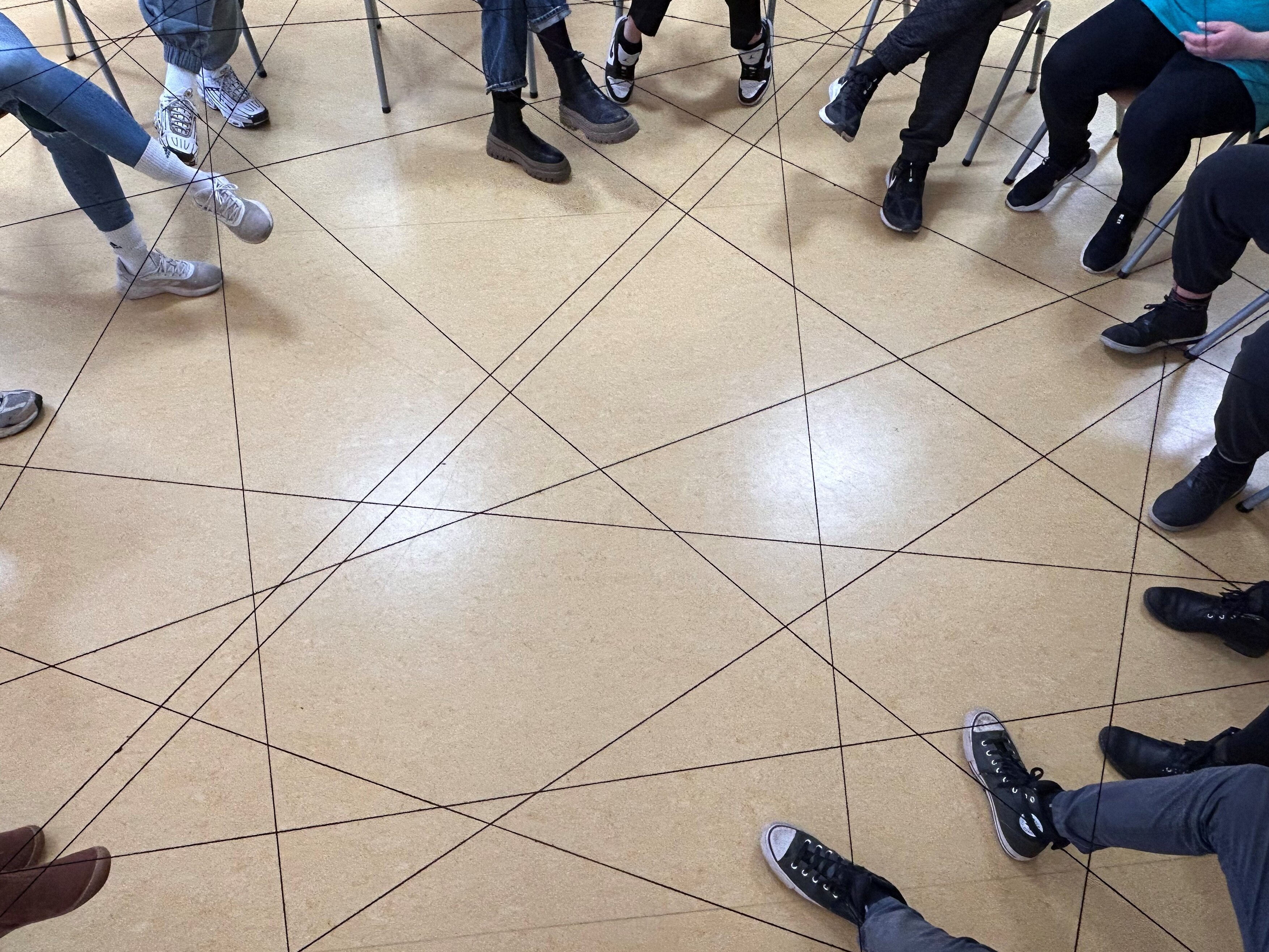Zu sehen ist ein Fadennetz, welches zwischen mehreren Personen in einem Stuhllreis gesponnen ist. Von den Personen sind nur Schuhe und Beine zu sehen.