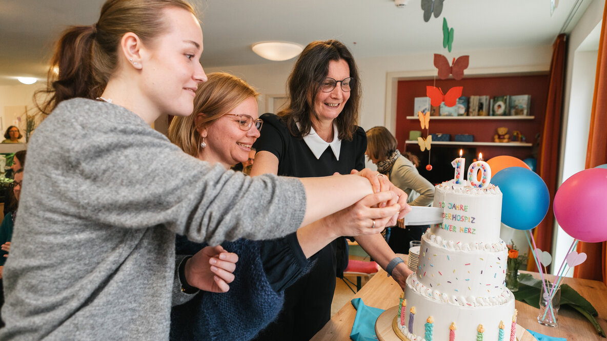 Drei Personen schneiden gemeinsam eine Torte an, die auf einem Tisch steht.