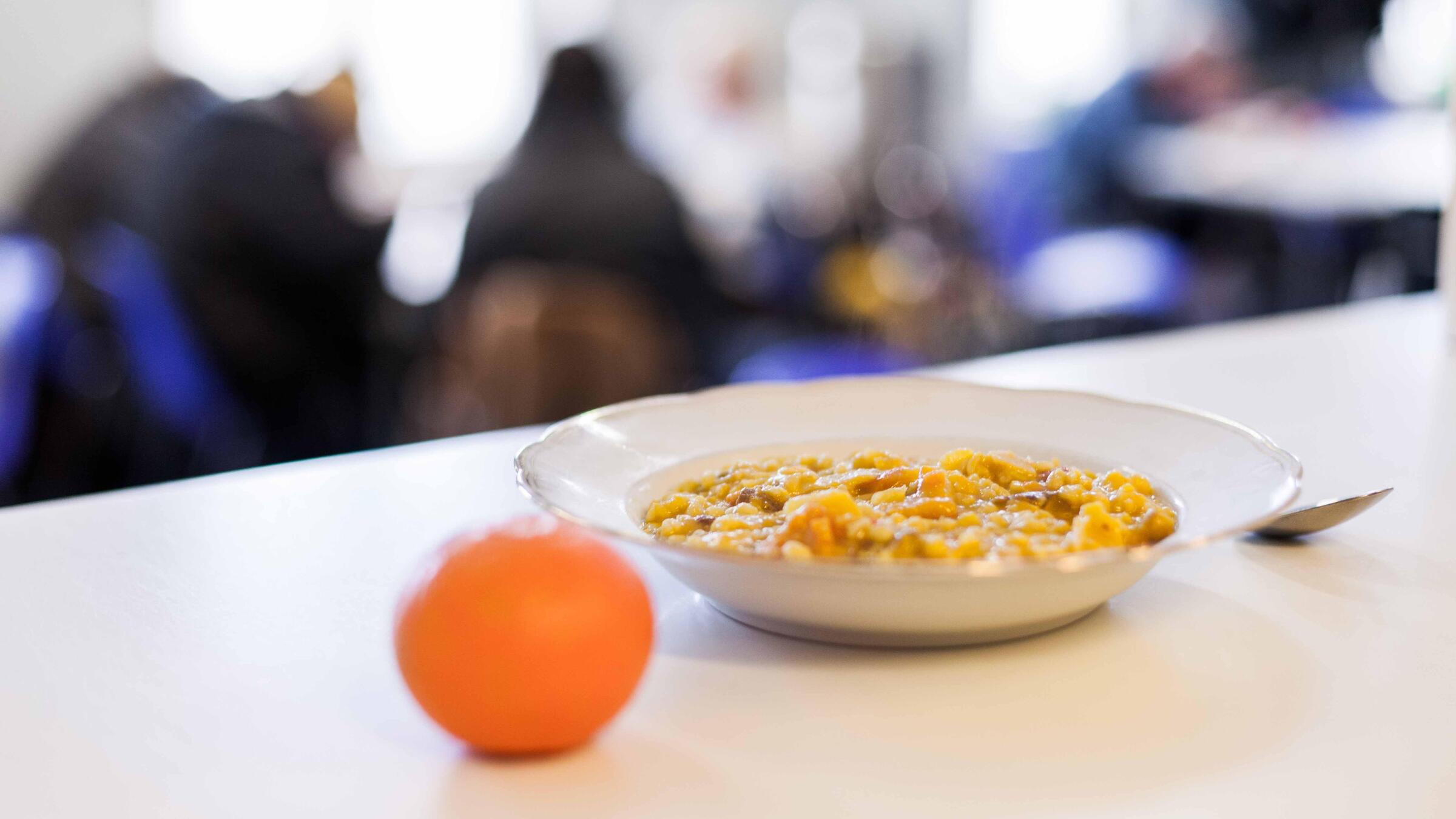 Auf einem weißen Tisch steht ein mit warmen Essen gefüllter Teller. Daneben liegt eine Mandarine und ein Löffel.