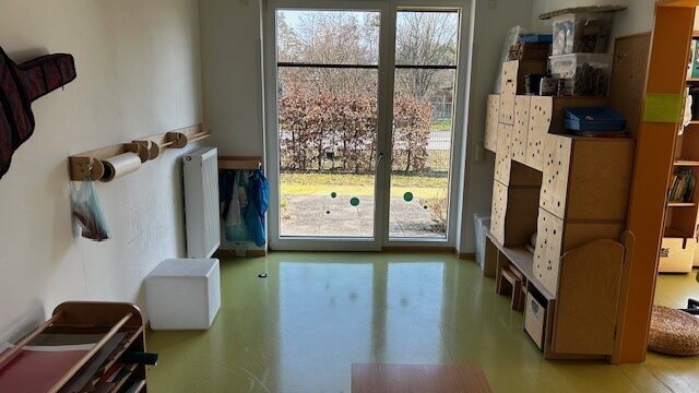 In einem Raum mit hoher Fensterfront stehen Regale mit Materialien und Utensilien für Kreatives sowie ein Tisch und Sitzmöbel.