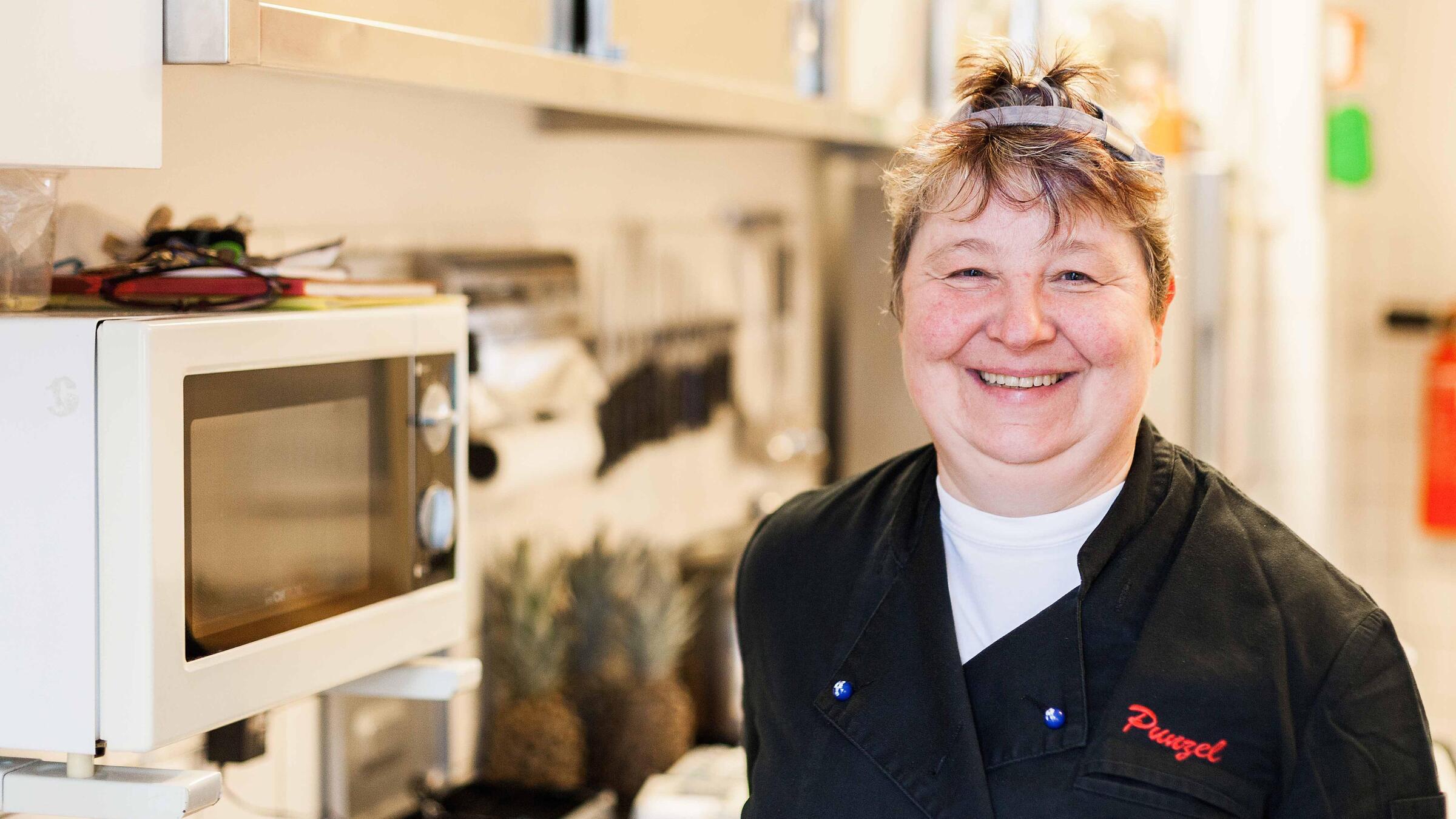 Eine Frau in einer schwarzen Kochjacke lächelt in die Kamera. Sie steht in einer Küche, neben ihr ist eine Mikrowelle und eine Küchenzeile zu erkennen.
