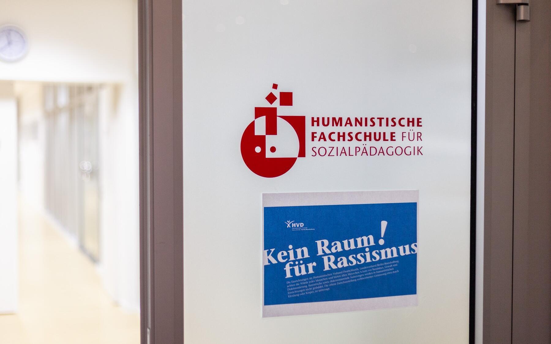 Auf einer Tür im Inneren des Fachschulgebäudes ist das Schullogo zu sehen. Darunter klebt ein DIN-A4-Blatt auf dem in Weiß auf Blau als Überschrift „Kein Raum für Rassismus!“ plus die Erklärung dazu zu lesen sind.