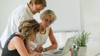 HospizKind Berlin: Hospizliche Versorgungs- und Beratungsstrukturen unterstützen und ausbauen Drei Frauen, zwei sitzend, sind an einem Schreibtisch und blicken auf ein Blatt Papier. Links davon steht ein Laptop.