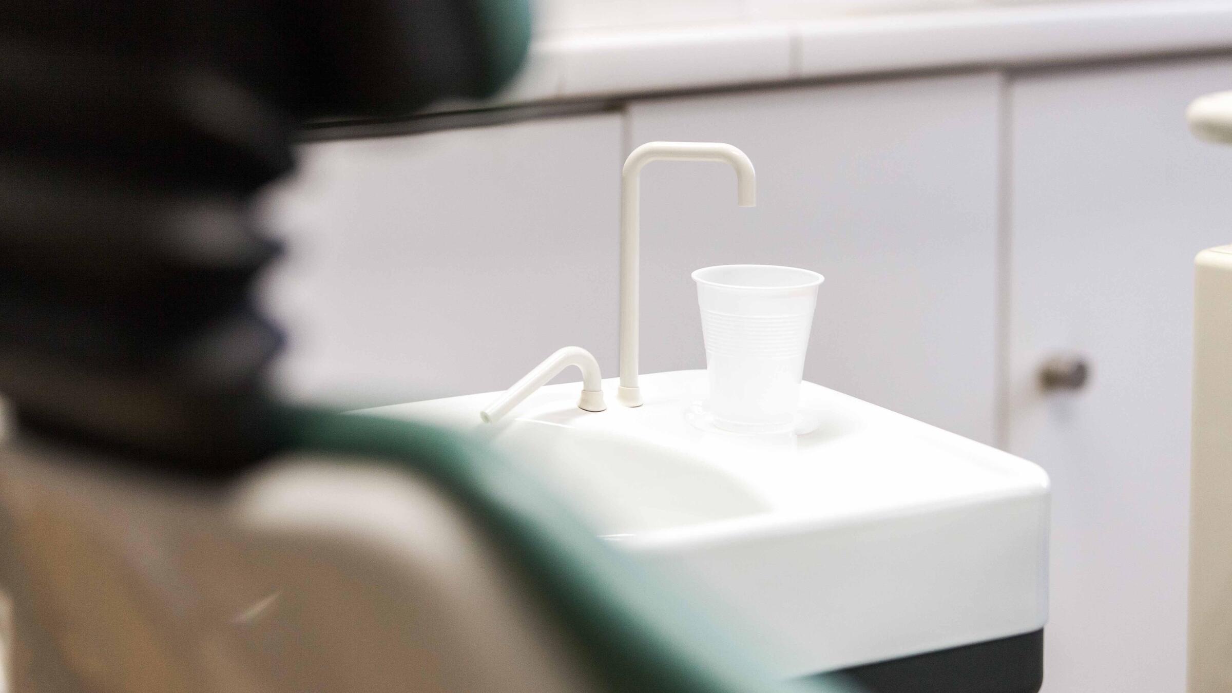 Verschwommen im Vordergrund ist angeschnitten ein Zahnarztstuhl zu erkennen. Im Zentrum sieht man ein kleines Waschbecken mit einem Plastikbecher, wie es in Zahnarztpraxen üblich ist.