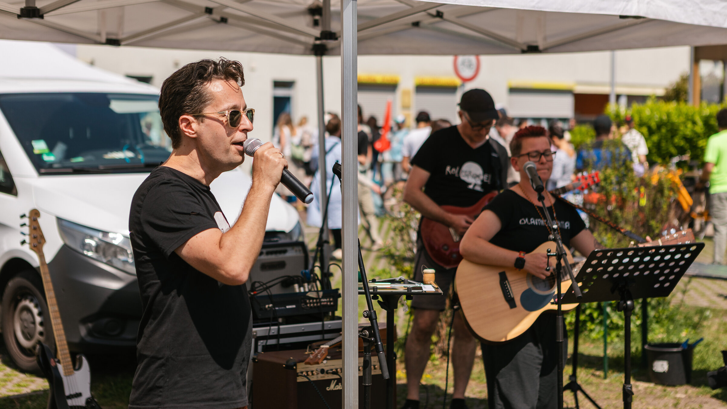 Eine Person im Vordergrund sing in eine Hintergrund. Im Hintergrund sieht man zwei weitere Erwachsene die Gitarre spielen.