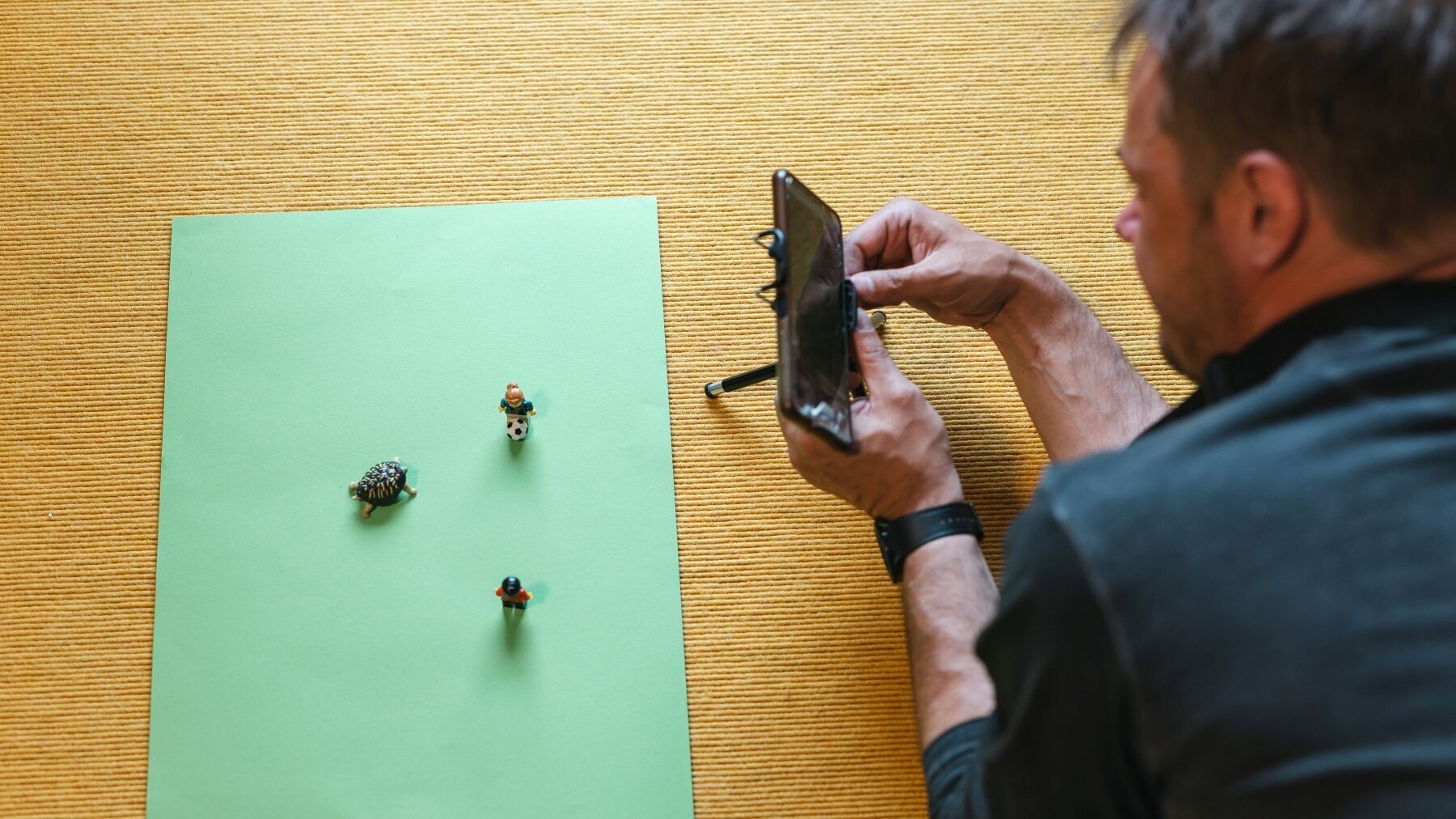 Ein Mann liegt auf einem Teppichboden und filmt mit einem Smartphone, das auf einem Ministänder steht, folgende Szenerie: Auf einem grünen Blatt stehen eine Minischildkrötenfigur und zwei Legofiguren. Vor einer Figur liegt ein Minifußball.