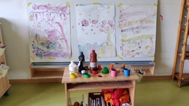 Ein Regal auf Rollen mit Kreativuntensilien steht in einem Raum. Dahinter an der Wand sind an einer weißen Tafel mit Klebestreifen drei große Bilder befestigt, die von Kindern gemalt wurden.