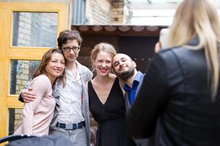 Humanistische Lebensfeiern in Berlin und Brandenburg Vier Menschen in festlicher Kleidung stehen nebeneinander im Freien und werden von einer Frau mit dem Handy fotografiert.