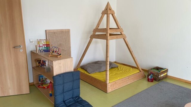 u sehen sind ein Teppich, ein Regal mit Spielzeug und eine Pyramidenkonstruktion aus Holz, auf deren Grundfläche eine Matte und ein Kissen liegen.