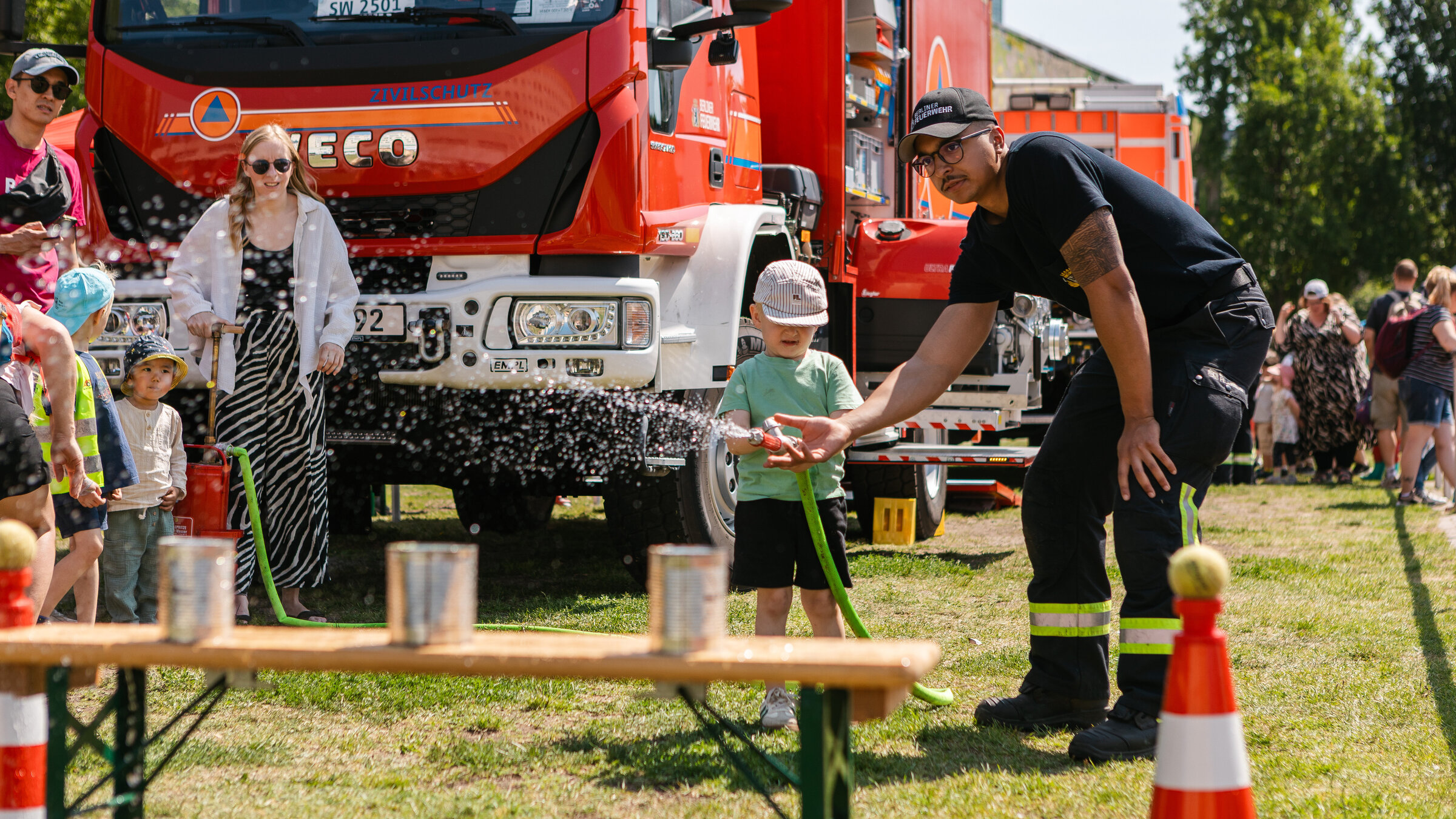 Ein kleiner junge hält einen dicken Wasserschlauch aus dem es sprüht. Ein Feuerwehrmann steht neben ihm gebäugt und hilfe ihm. Im Hintergrund ist ein großes Feuerwehrauto zu sehen.