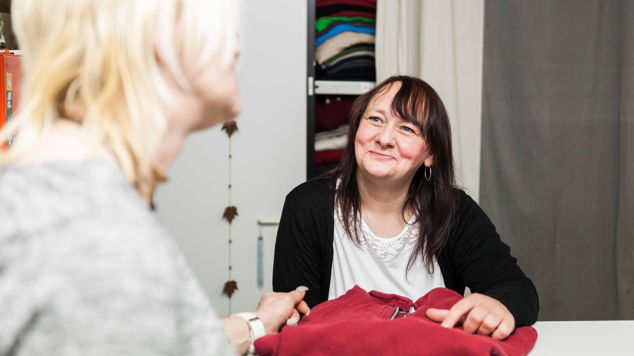 Zu sehen ist eine Frau in einer schwarzen Jacke, die sich über eine Art Theke lehnt. Vor ihr liegt ein roter Pullover, den sie gerade an eine andere Frau vor der Theke aushändigt. Hinter ihr ist ein Schrank mit mehr Kleidung zu erkennen.