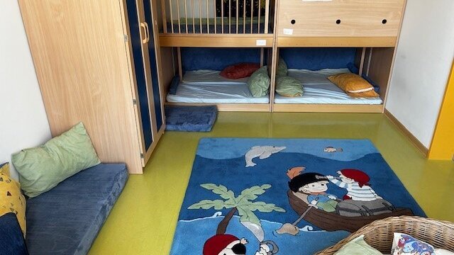 In einem Raum liegt ein Teppich mit Kinderpiratenmotiv. Hinter an der Wand gibt es offene Schrankteile mit Matratzen und Kissen, links davor steht ein Kleiderschrank. Unten im Bild sind auch Kissen an der Wand und in einem Korb zu sehen.