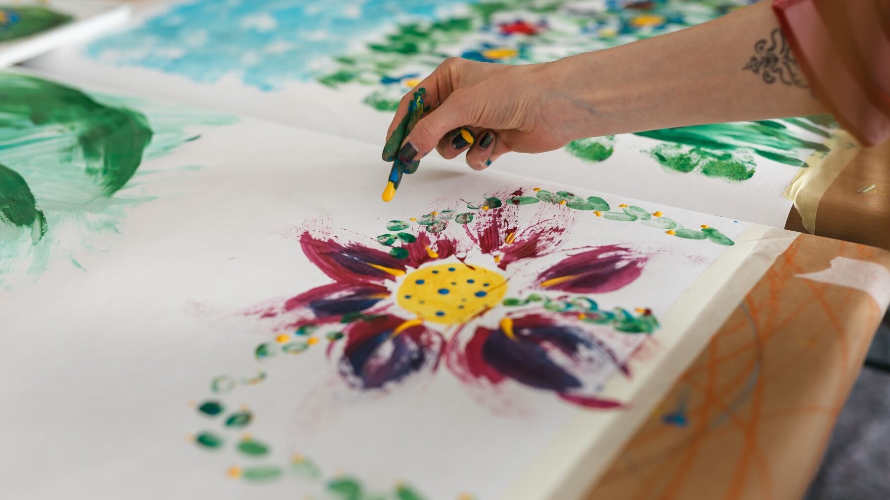Eine rechte Hand oben im Bild malt eine Blüte auf einer weißen Leinwand, die auf einem Tisch liegt.