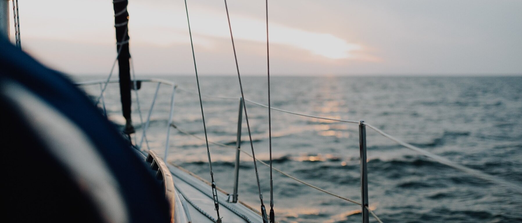 Links im Bild ist der rechte vordere Bereich einer Segeljacht und rechts sowie hinten sind ein Gewässer und der Horizont zu sehen.
