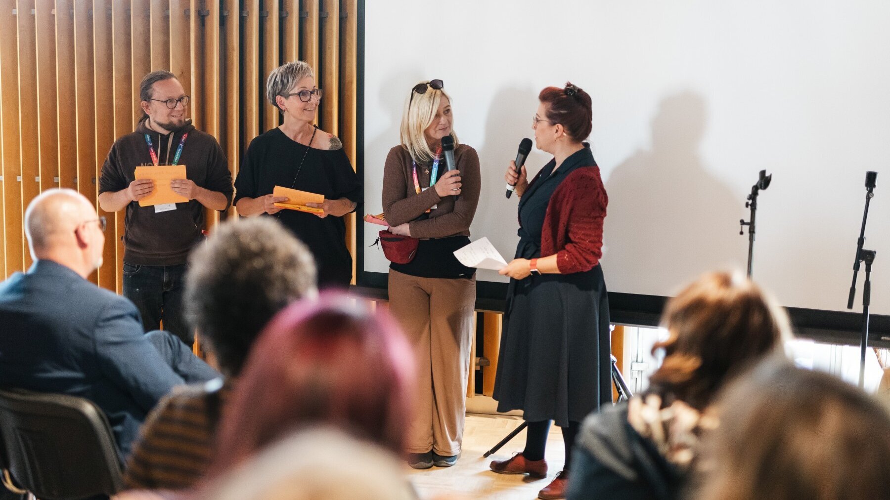 Ein Mann und drei Frauen stehen nebeneinander, die Frau rechts spricht in ein Mikrofon. Im Vordergrund sind von hinten sitzende Personen zu sehen.