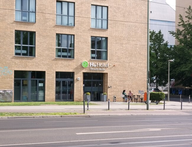 Zu sehen ist eine Hausecke mit großen Fenstern an einer großen Straße. Die Eingagstür ist mit H+ Hotel 4Youth Berlin beschriftet.