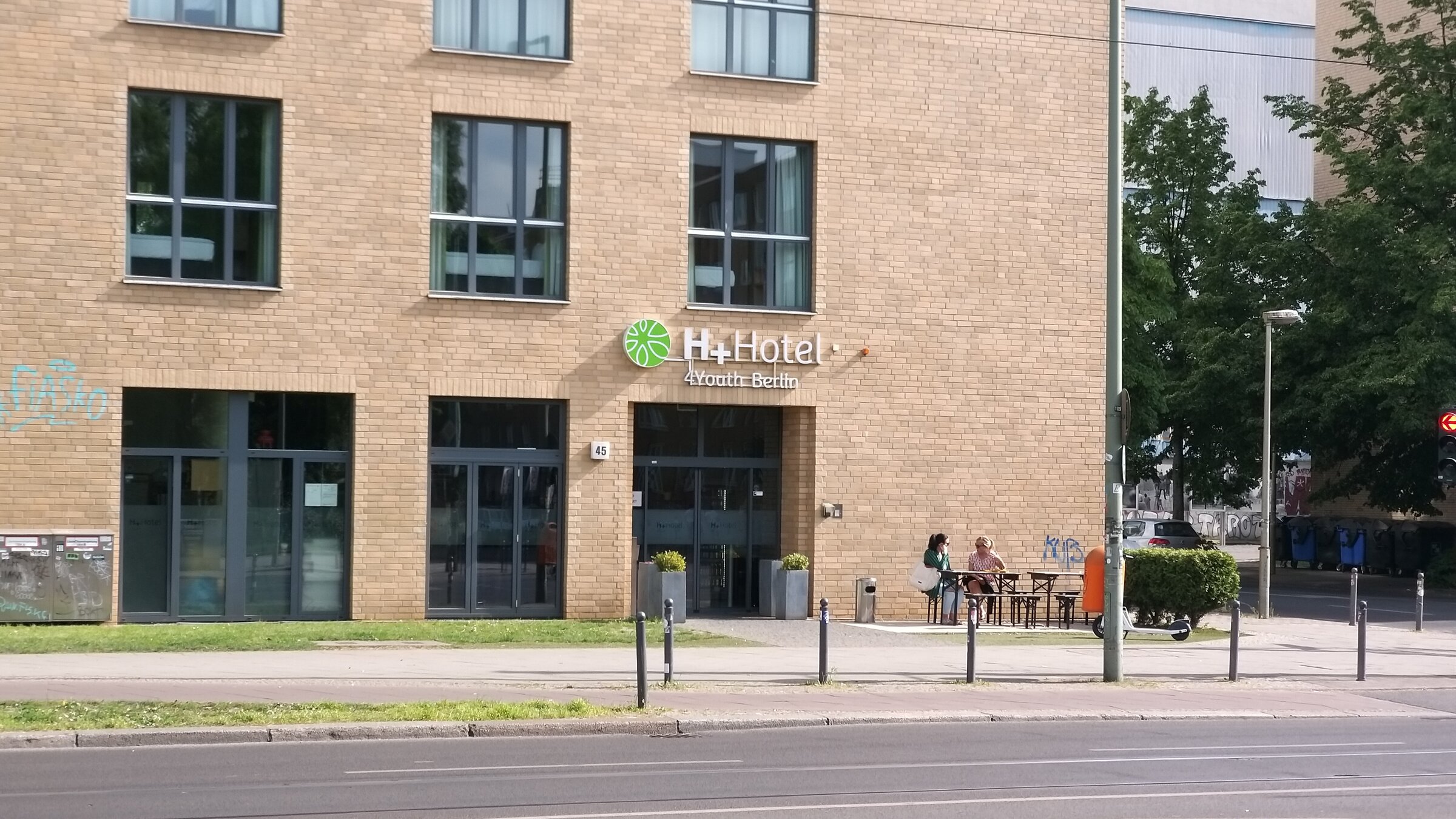 Zu sehen ist eine Hausecke mit großen Fenstern an einer großen Straße. Die Eingagstür ist mit H+ Hotel 4Youth Berlin beschriftet.