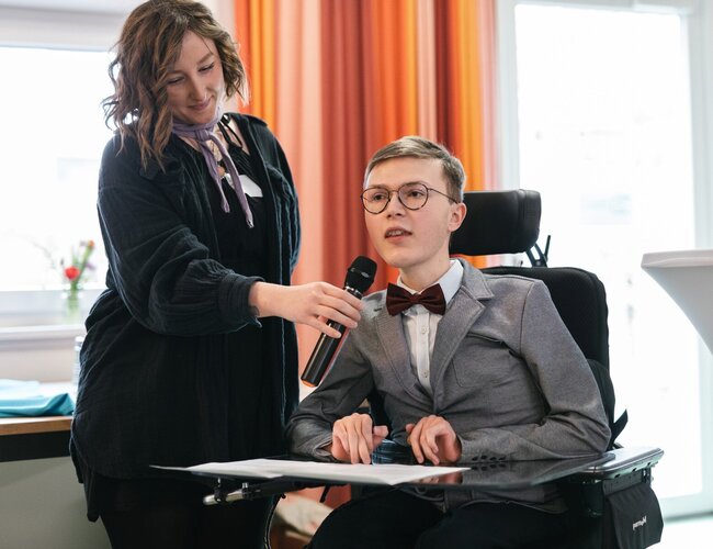 Lorenz Klar sitzt im Rollstuhl. Links daneben steht eine Frau, die mit ihrer rechten Hand ein Mikrofon vor seinen Mund hält, in das er spricht.