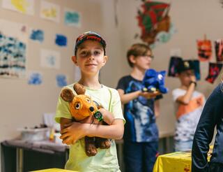 Kinderhospiz Berliner Herz: Geschwisterangebote Ein Junge mit Basecap umarmt ein Mauskuscheltier und lächelt. Rechts im Hintergrund sind verschwommen weitere Kinder zu sehen.