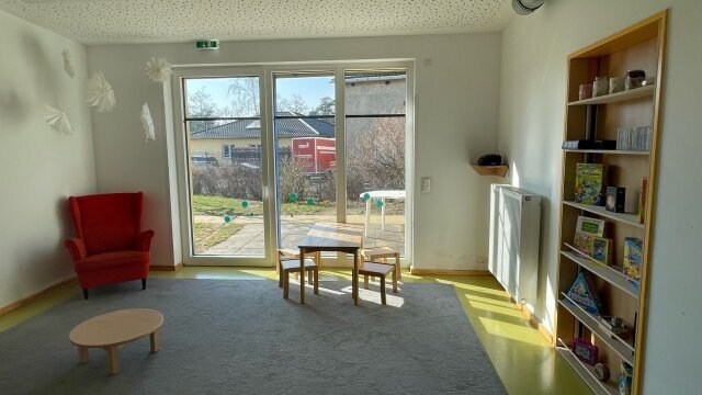 In einem Raum mit hoher Fensterfront liegt ein Teppich. Daruf stehen ein flacher Tisch, ein Sessel und eine Sitzecke mit vier Hockern. Rechts in der Wand ist eine Regal eingebaut, auf dem Spielzeug steht.