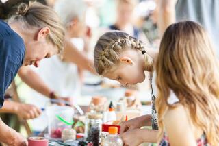 Pressefotos Engagement Eine Frau links und zwei Mädchen rechts basteln etwas an einem Tisch im Freien bei sommerlichem Wetter.