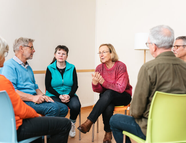 Zu sehen ist eine Gesprächsrunde von sechs Personen, die auf Stühlen in einem Kreis sitzen. Im Zentrum sitzen zwei Frauen. Die Frau rechts trägt einen roten Pulli und schein gerade zu sprechen. Sie hat den Mund leicht geöffnet und ist nach vorne gebeugt.