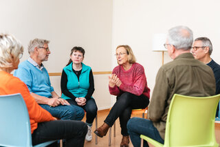 Kontaktstelle PflegeEngagement: Unterstützung für pflegende Angehörige Zu sehen ist eine Gesprächsrunde von sechs Personen, die auf Stühlen in einem Kreis sitzen. Im Zentrum sitzen zwei Frauen. Die Frau rechts trägt einen roten Pulli und schein gerade zu sprechen. Sie hat den Mund leicht geöffnet und ist nach vorne gebeugt.