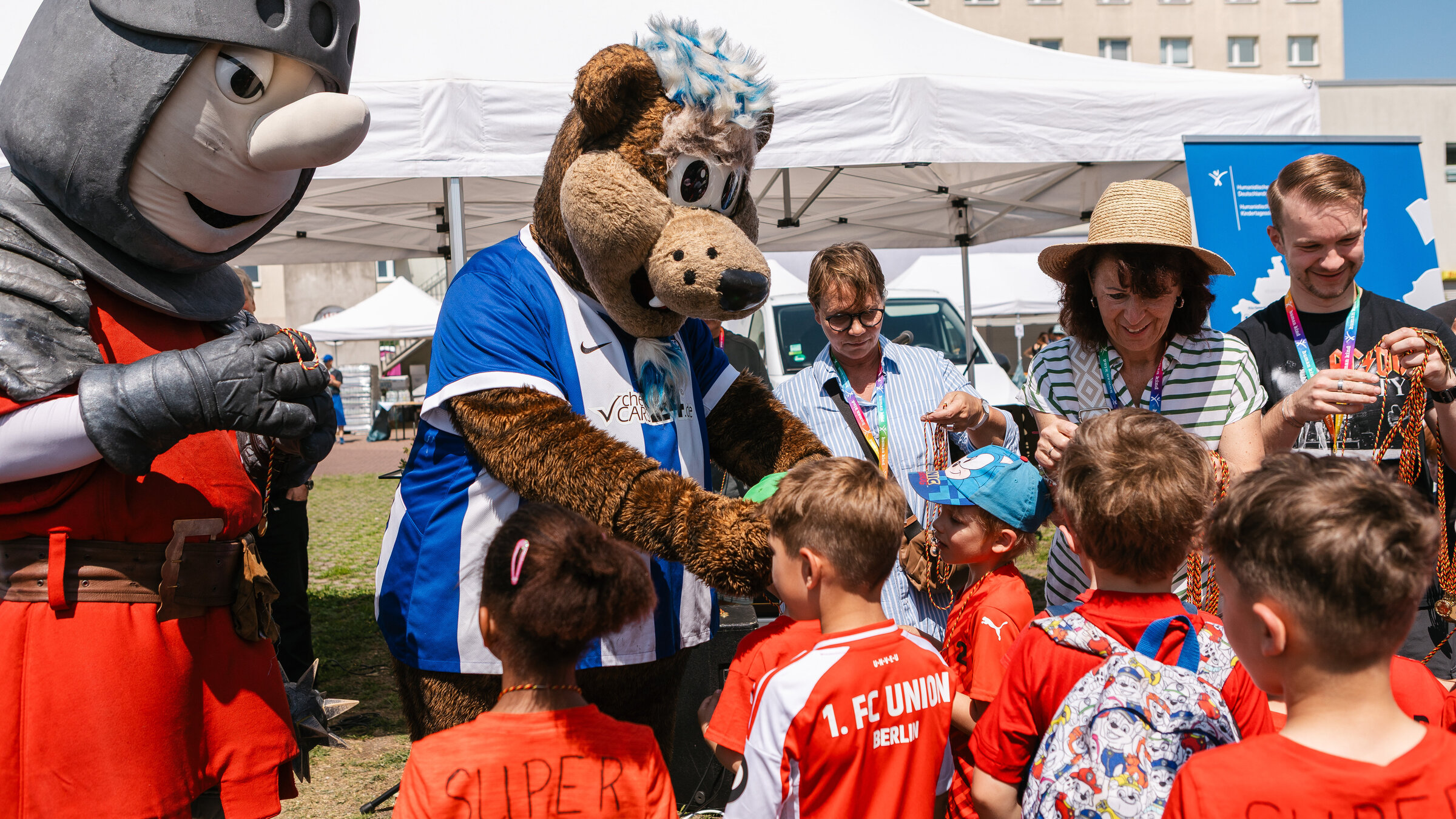 Die Maskottchen Ritter Keule und Herthinho stehen vor einer Gruppe kleiner Kinder in Fußballtrikots und verteilen Medaillen.