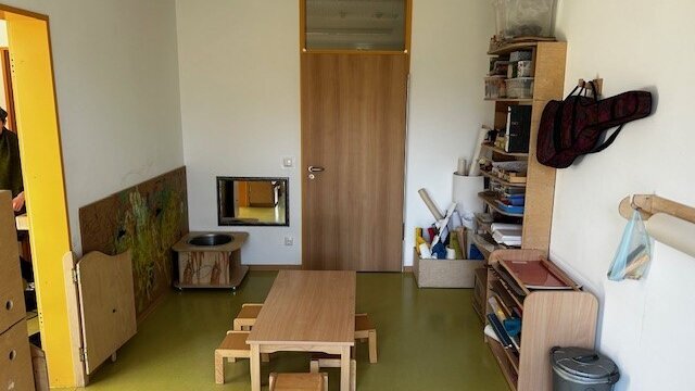 In einem Raum stehen Regale mit Materialien und Utensilien für Kreatives sowie ein Tisch und Hocker. Rechts an der Wand hängt eine Gitarrentasche.