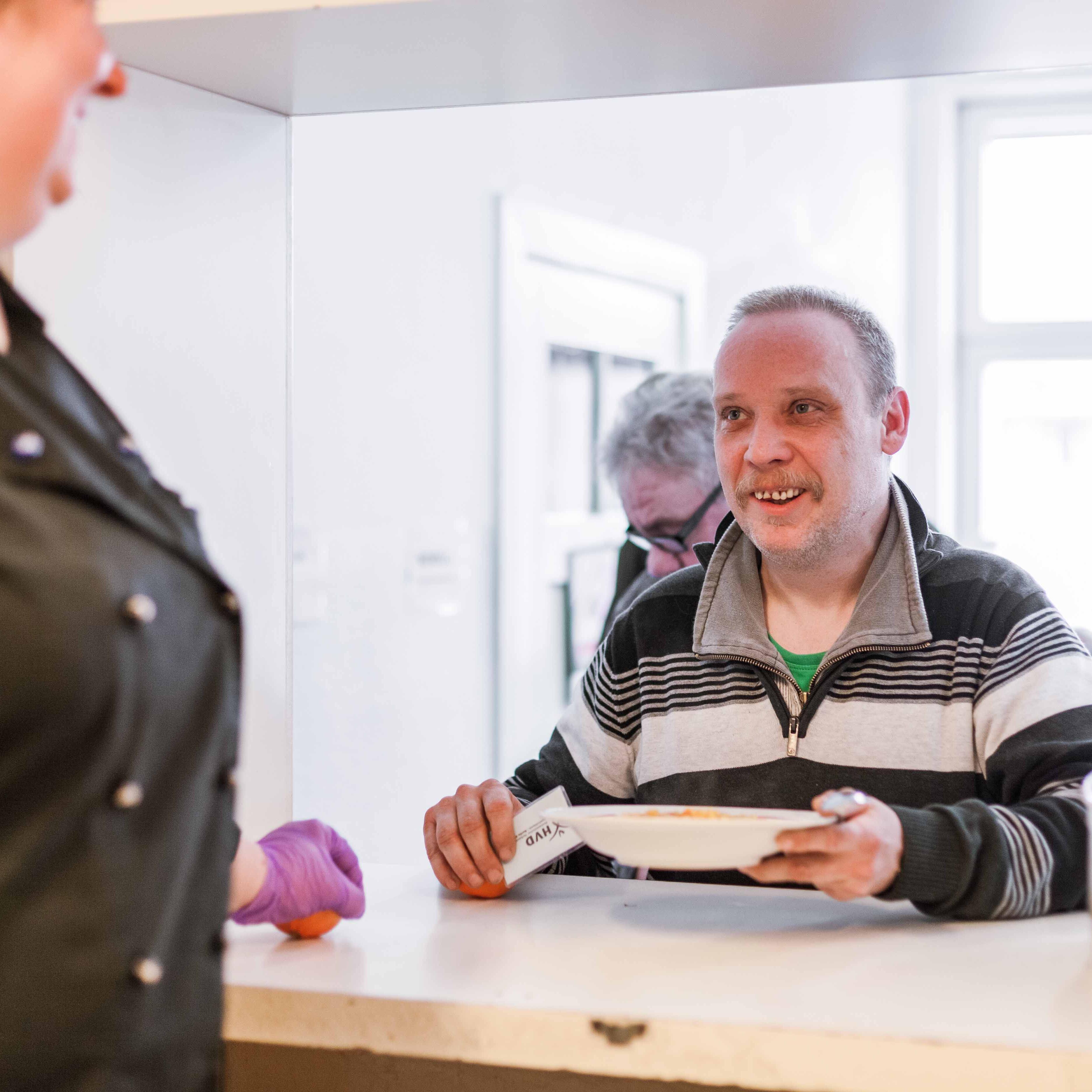 Links steht angereissen eine Frau in einer schwarzen Kochjacke und schaut druch die Luke der Essensausgabe auf einem Mann, der sich gerade einen Teller mit warmen Essen und eine Mandarine von der Theke nimmt. Sie lächeln sich an. 