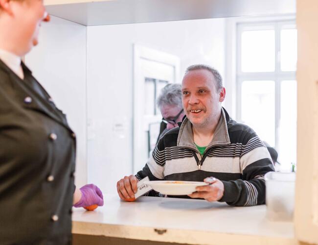 Links steht angereissen eine Frau in einer schwarzen Kochjacke und schaut druch die Luke der Essensausgabe auf einem Mann, der sich gerade einen Teller mit warmen Essen und eine Mandarine von der Theke nimmt. Sie lächeln sich an. 