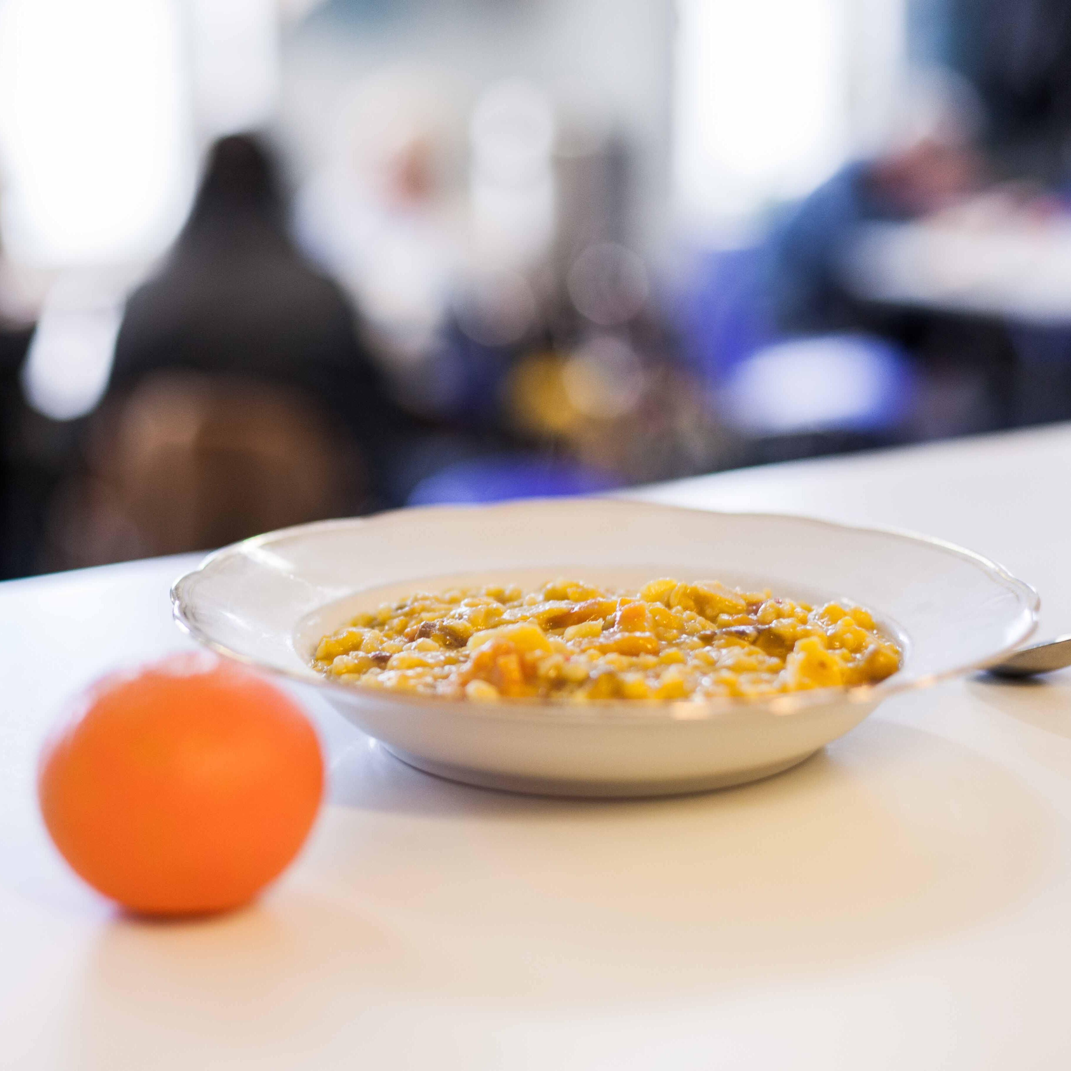 Auf einem weißen Tisch steht ein mit warmen Essen gefüllter Teller. Daneben liegt eine Mandarine und ein Löffel.