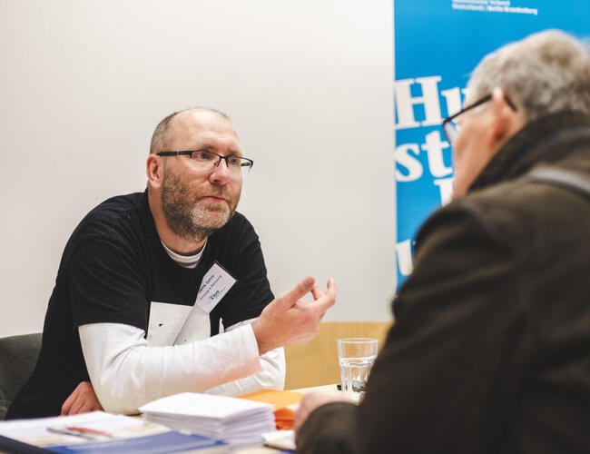 Jens Gehre, Nachlassberater beim Humanistischen Verband Berlin-Brandenburg, sitzt an einem Tisch und berät einen Mann, der ihm gegenüber am Tisch sitzt.