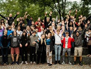 JuHus Brandenburg Gruppenbild Eine Gruppe mit mehr als 20 jungen Menschen steht teilweise erhöht auf einer Steintreppe vor grünen Bäumen. Sie schauen in die Kamera, lächeln und ein paar halten ihre Hände in die Luft.