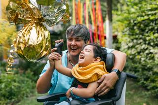 Kinderhospiz Berliner Herz Eine Betreuerin umarmt ein schwer erkranktes Kind mit körperlichen Beeinträchtigungen im Garten vom Kinderhospiz Berliner Herz. Sie hält mit ihrer rechten Hand die rechte Hand des Kindes, um auf eine Piñata aus goldglänzender Folie zu schlagen. Beide lachen.