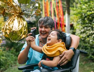 Kinderhospiz Berliner Herz Eine Betreuerin umarmt ein schwer erkranktes Kind mit körperlichen Beeinträchtigungen im Garten vom Kinderhospiz Berliner Herz. Sie hält mit ihrer rechten Hand die rechte Hand des Kindes, um auf eine Piñata aus goldglänzender Folie zu schlagen. Beide lachen.